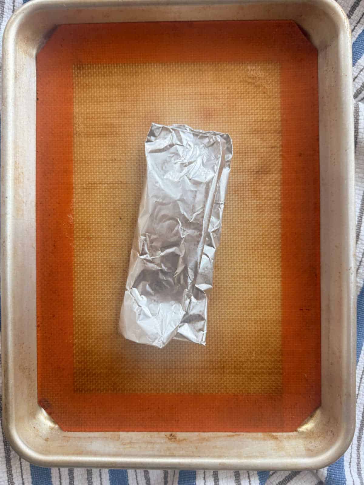 A foil-wrapped package sits on a silicone baking mat placed on a rimmed metal baking sheet.