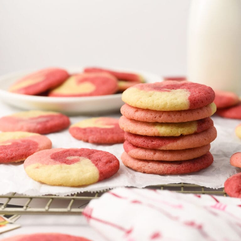 A stack of pink and white Valentine's swirl cookies sits on parchment paper, with more cookies and a bottle of milk in the background.