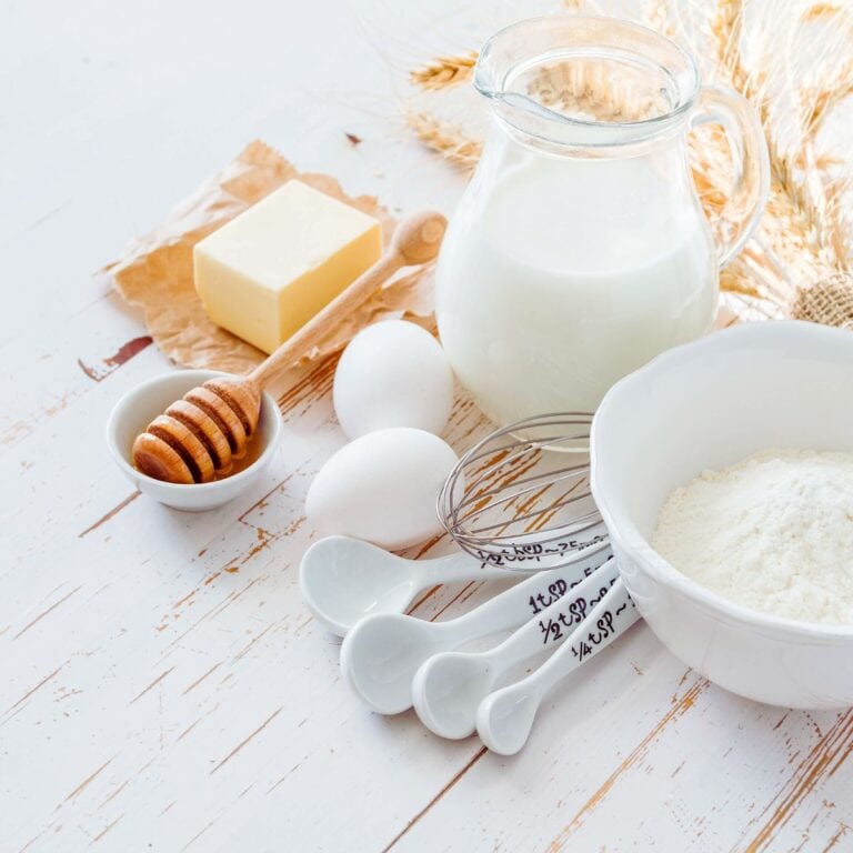 A bowl of flour, eggs, butter, milk, honey, and measuring spoons are arranged on a white wooden surface with wheat stalks in the background.