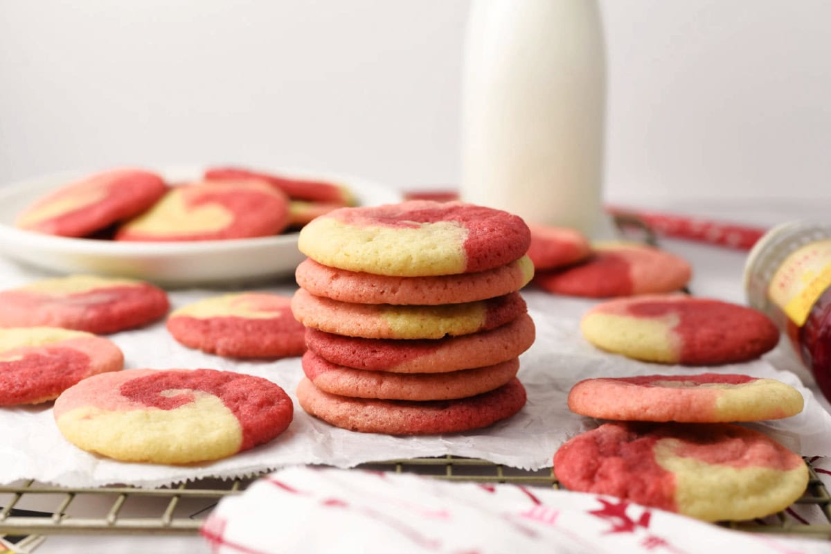 A stack of red and white swirled cookies sits on a cooling rack, surrounded by more cookies, with a bottle of milk and a plate of cookies in the background.