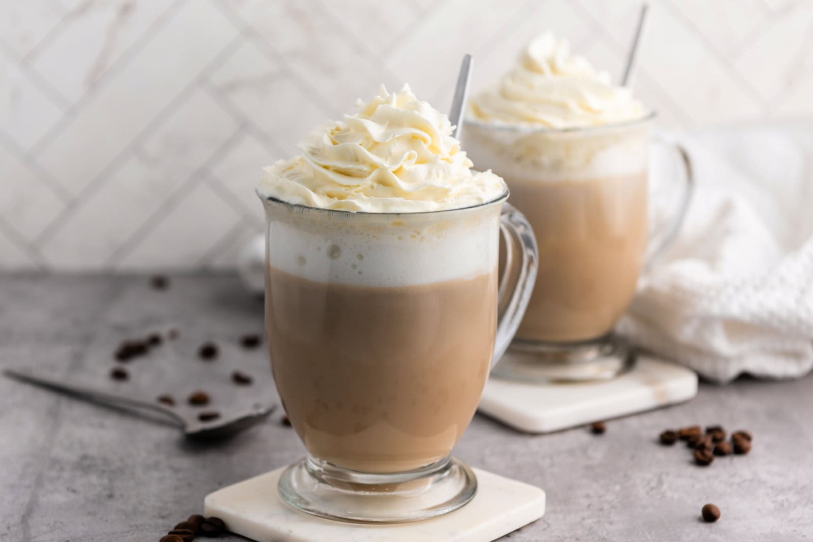 Two glass mugs of coffee topped with whipped cream sit on coasters, with scattered coffee beans and a spoon on a grey surface.