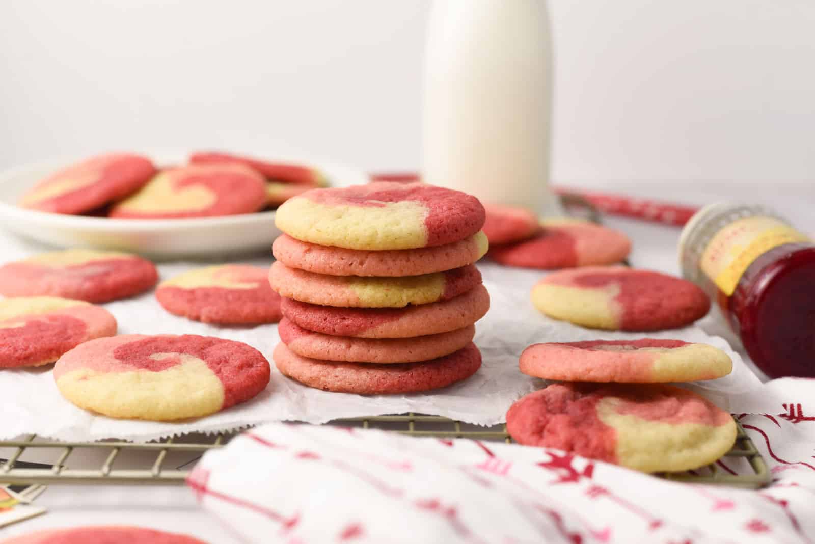 A stack of red and yellow marbled cookies is surrounded by more cookies, a bottle of milk, and a jar of jam on a wire rack and parchment paper.