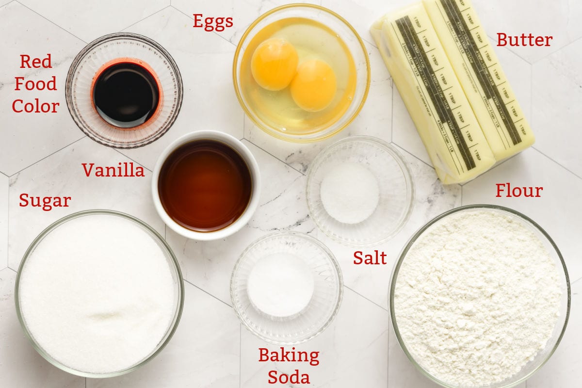 Various baking ingredients in bowls and containers, labeled as eggs, butter, flour, salt, baking soda, sugar, vanilla, and red food color, arranged on a white countertop.