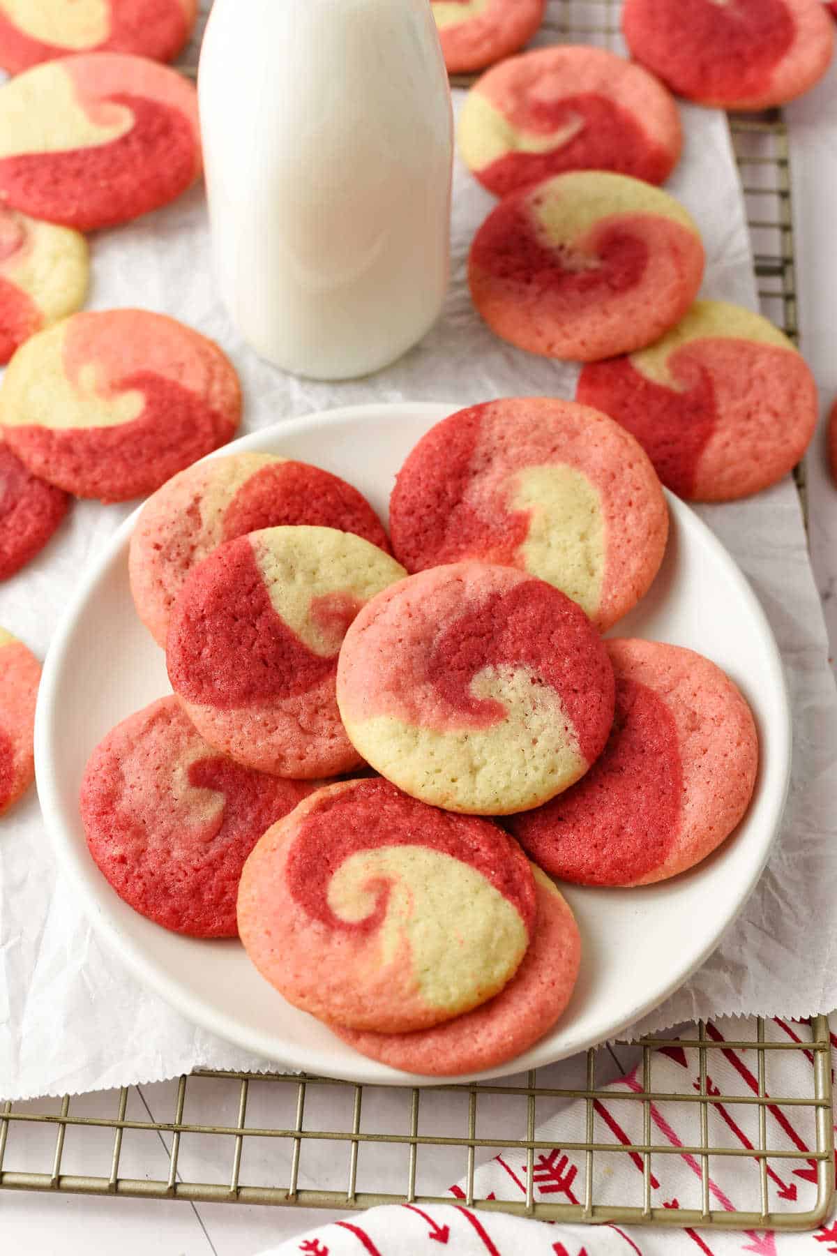 A plate of red, pink, and white swirl cookies sits beside a bottle of milk, with more cookies on a cooling rack in the background.