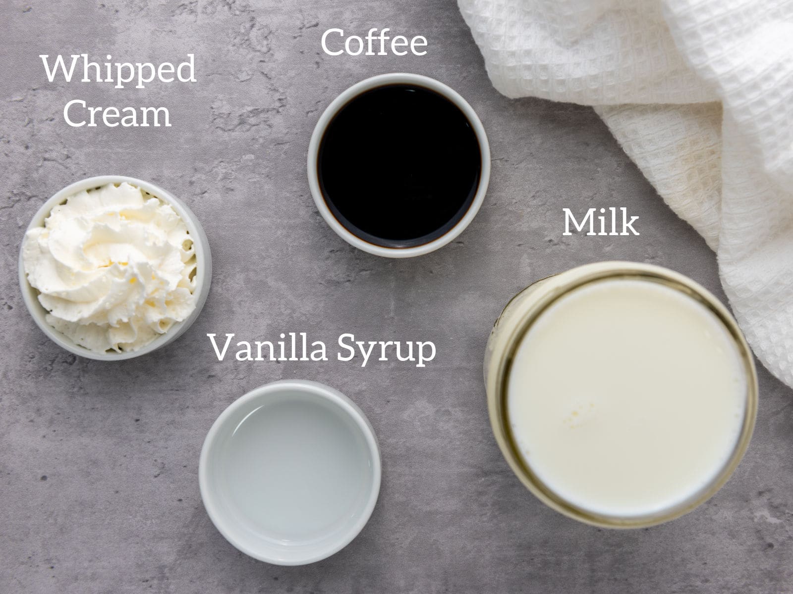 Four labeled ingredients on a gray surface: whipped cream, coffee, vanilla syrup, and milk, arranged in small bowls and a glass. A white textured cloth is in the top right corner.