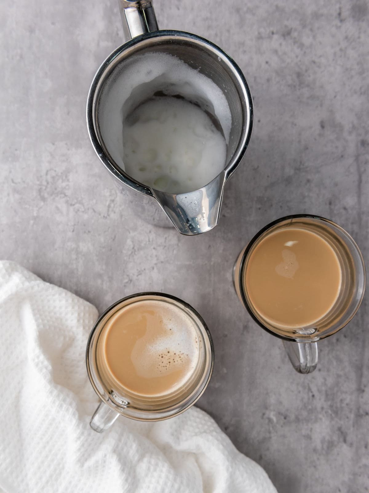 A metal milk frothing pitcher with steamed milk beside two glass mugs of coffee on a gray surface, next to a white textured cloth.