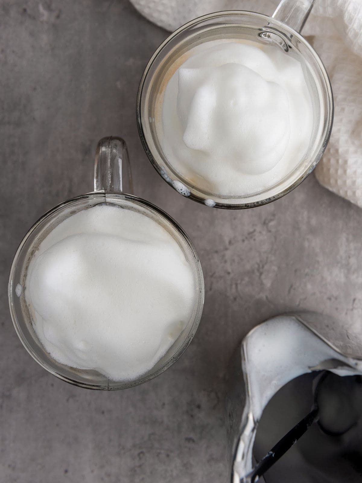 Two glass mugs filled with milk froth are placed on a gray surface, next to a metal frothing pitcher.