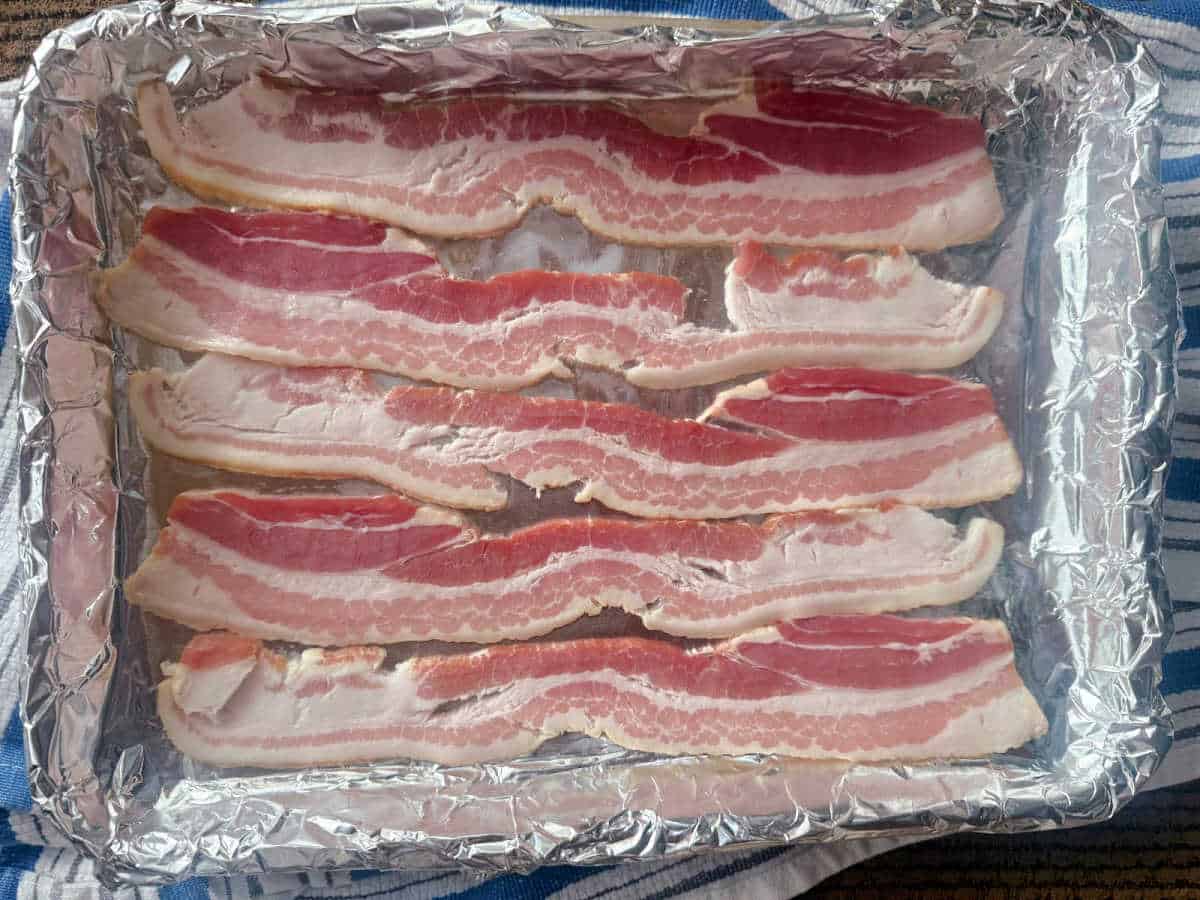 Raw bacon strips arranged on a foil-lined baking tray, ready to be cooked.