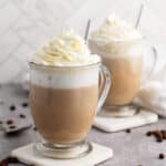 Two glass mugs of coffee topped with whipped cream are placed on coasters, with metal spoons inside, on a gray surface.