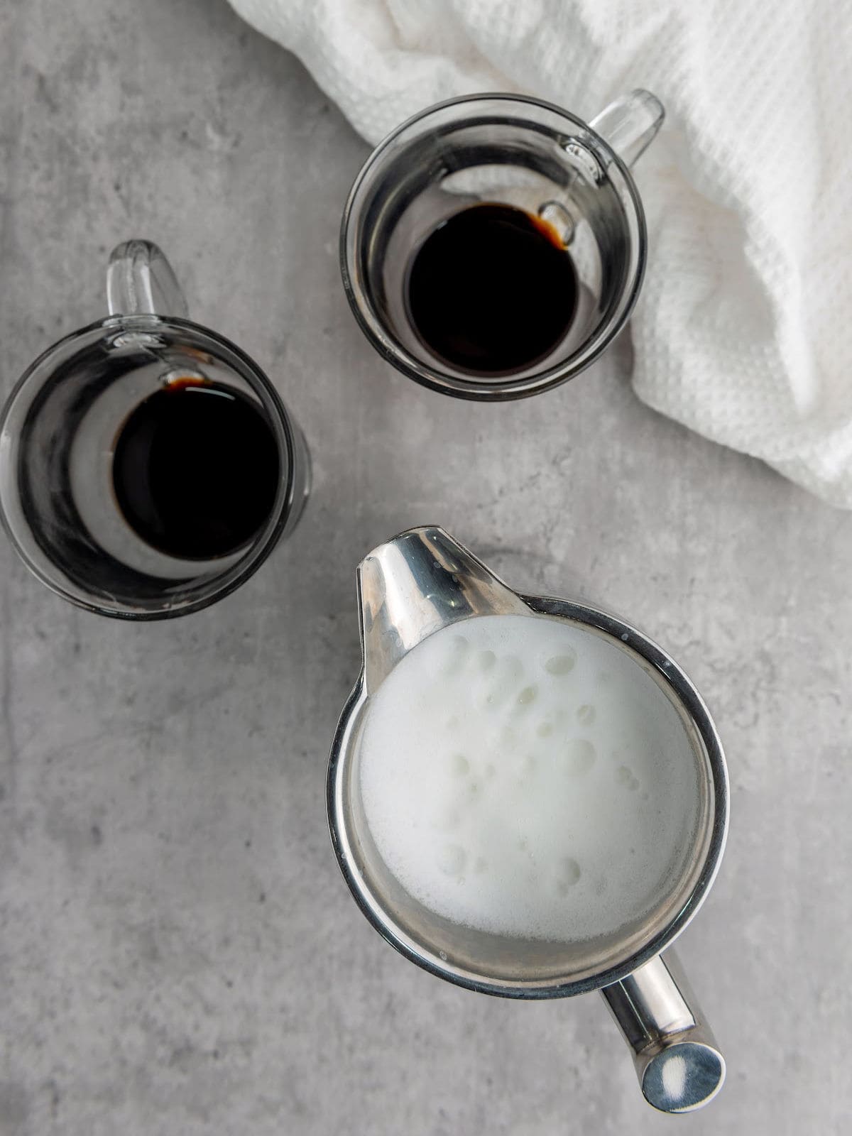 Two glass mugs with black coffee and a metal pitcher of steamed milk on a gray surface with a white cloth.