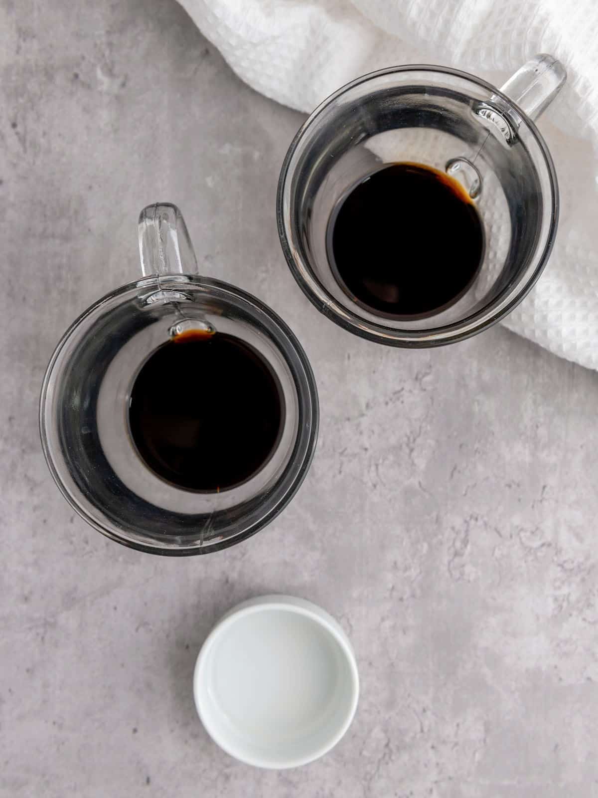 Two clear glass mugs filled with coffee, placed on a gray surface next to a small empty white dish and a white textured cloth.