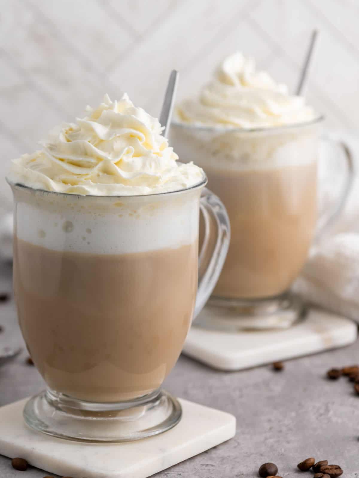 Two glass mugs filled with vanilla latte coffee topped with whipped cream, each on a coaster, with spoons inside. Whole coffee beans are scattered on the gray surface.