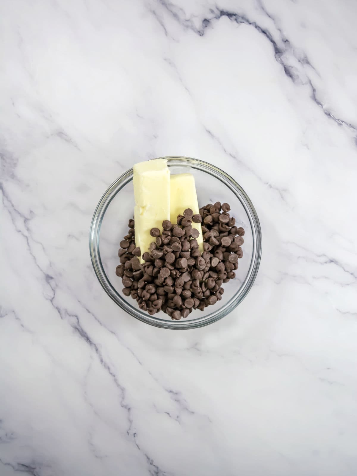 A glass bowl containing two sticks of butter and a pile of chocolate chips sits on a white marble surface.