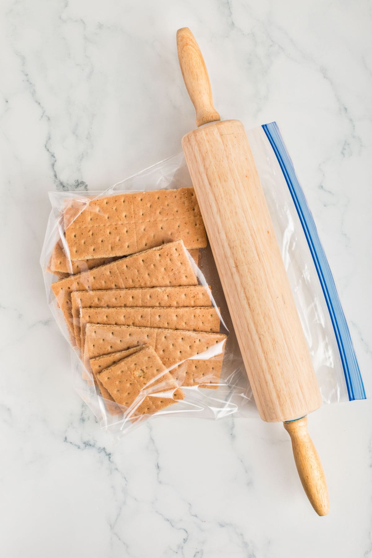 A rolling pin rests on a marble surface next to a resealable plastic bag filled with whole and broken graham crackers.