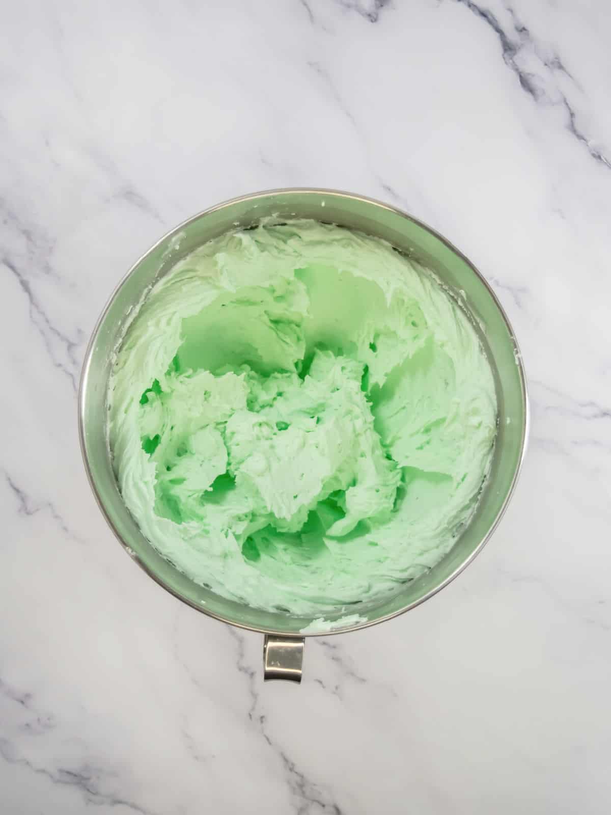 A metal mixing bowl filled with light green mint whipped frosting sits on a white marble surface.