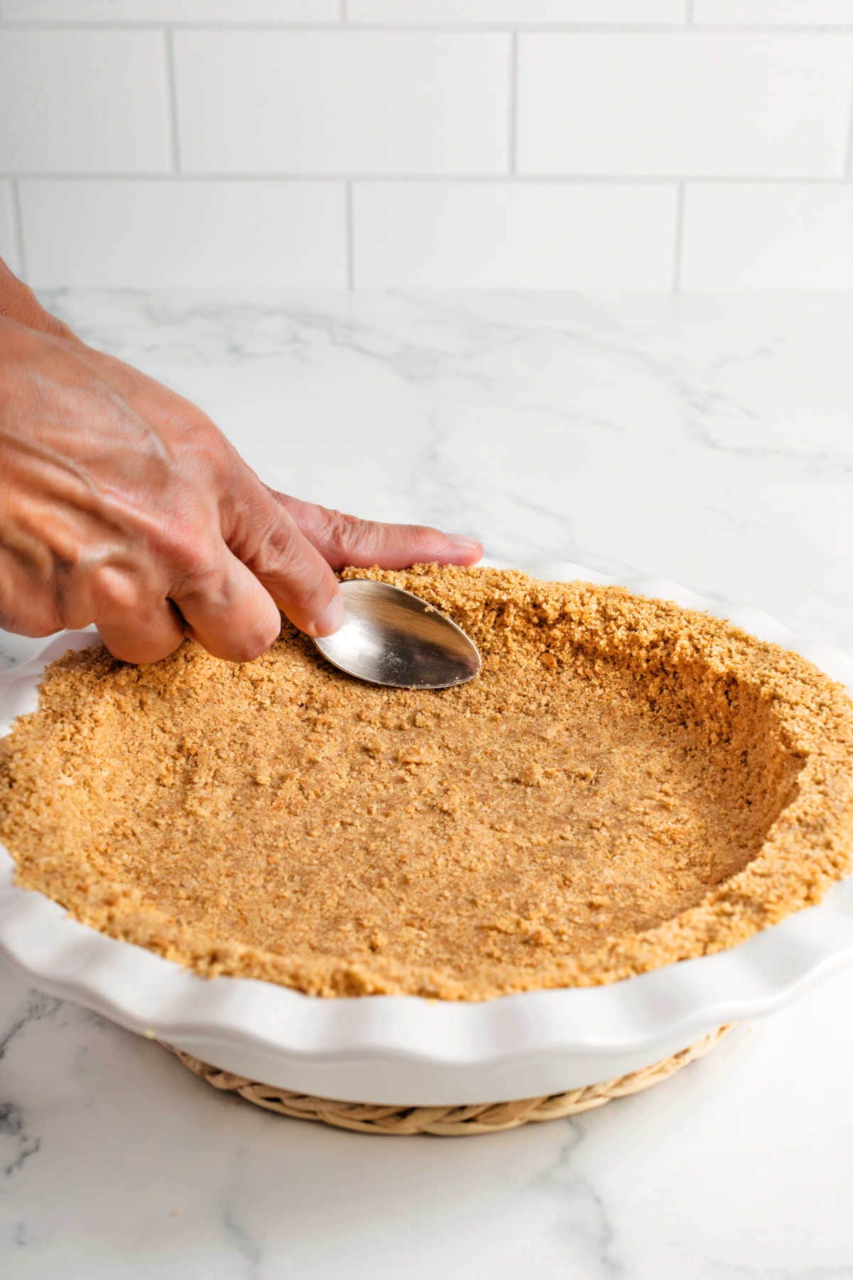 A hand presses a graham cracker crust into a pie dish using the back of a spoon on a marble countertop.