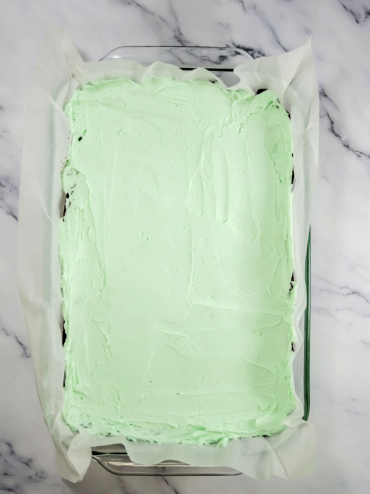 A rectangular glass baking dish lined with parchment paper and filled with a dessert topped with smooth, pale green frosting, set on a marble surface.