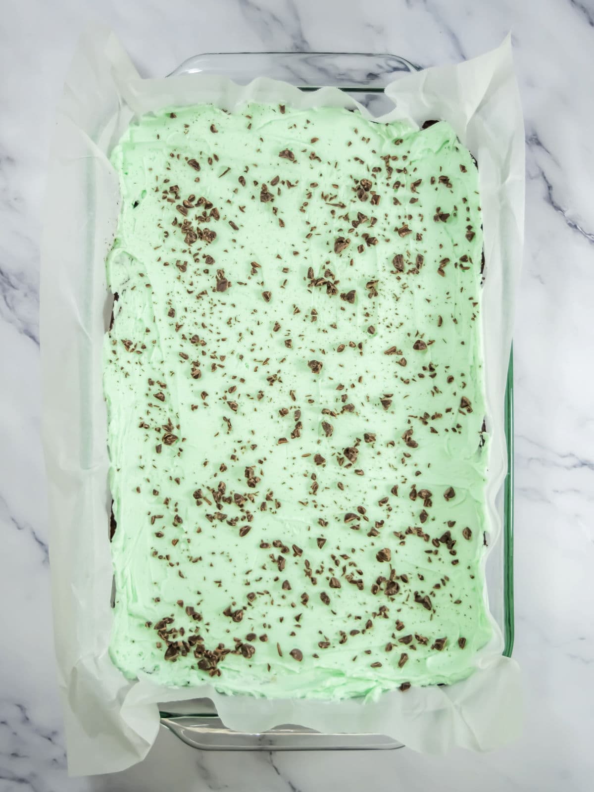 A rectangular pan filled with a dessert topped with light green frosting and sprinkled chocolate pieces, lined with parchment paper on a marble surface.