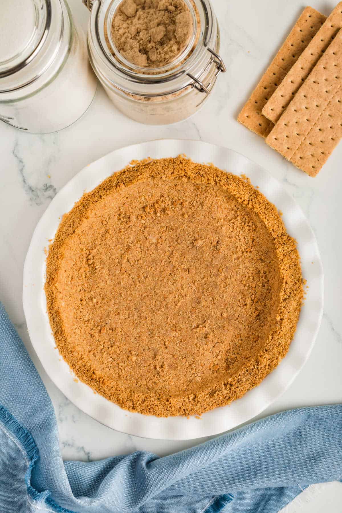 A pie dish filled with prepared graham cracker crust sits on a marble surface, with jars of sugar and brown sugar, graham crackers, and a blue cloth nearby.