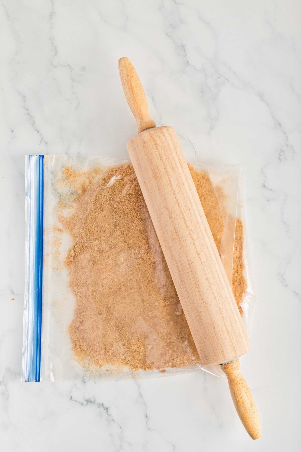A rolling pin is crushing graham crackers inside a sealed plastic bag on a marble countertop.