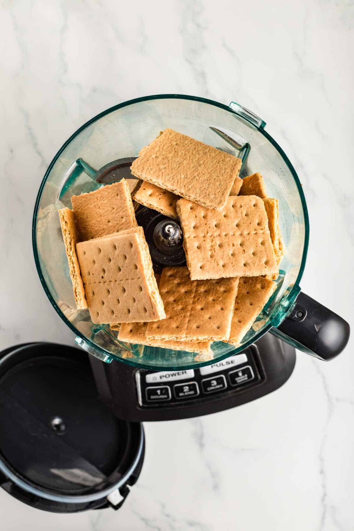 A food processor containing several graham crackers on a marble countertop with the lid removed.