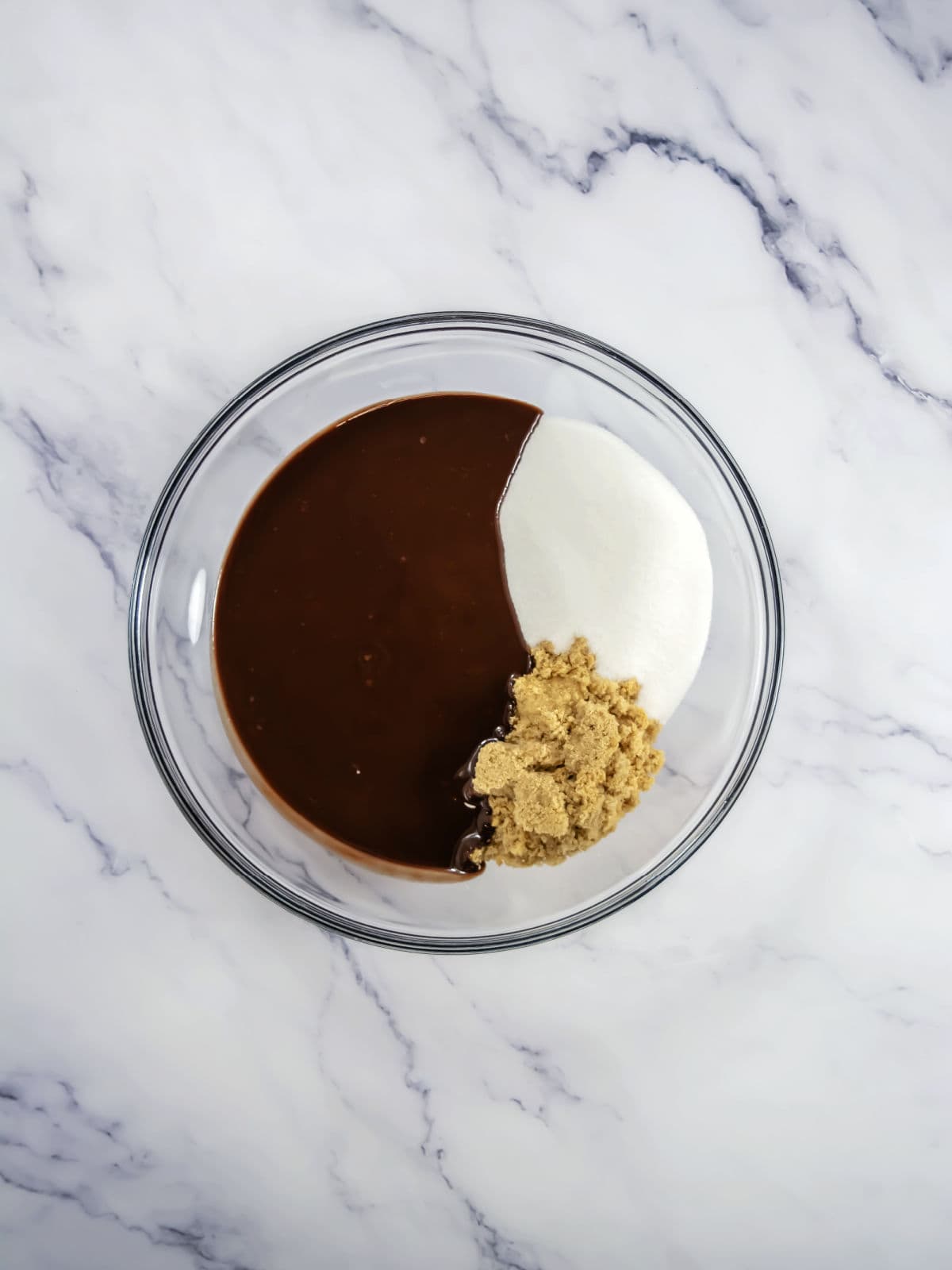 A glass bowl containing chocolate mixture, white granulated sugar, and brown sugar on a white marble surface.