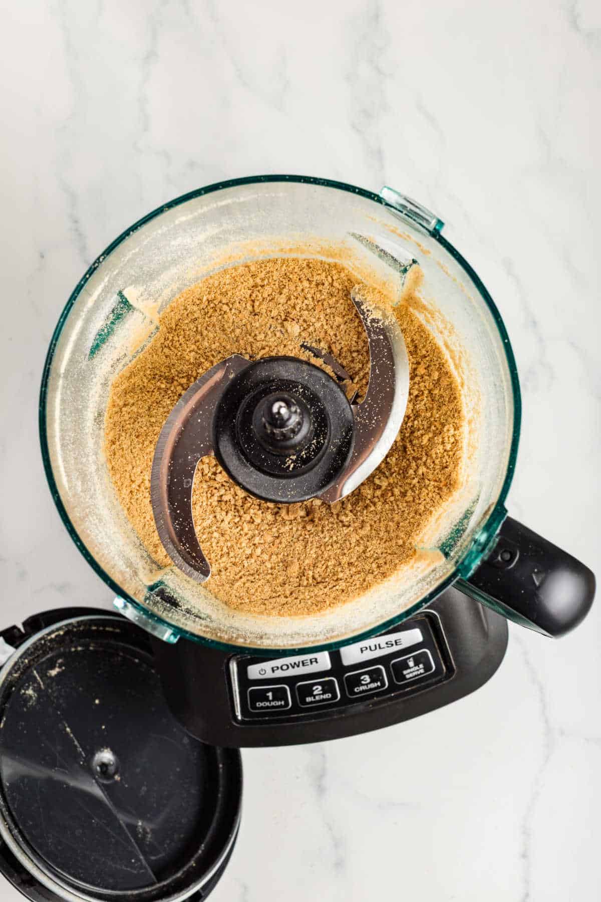 A food processor with crushed graham crackers inside, viewed from above on a marble surface. The lid is open and resting nearby.