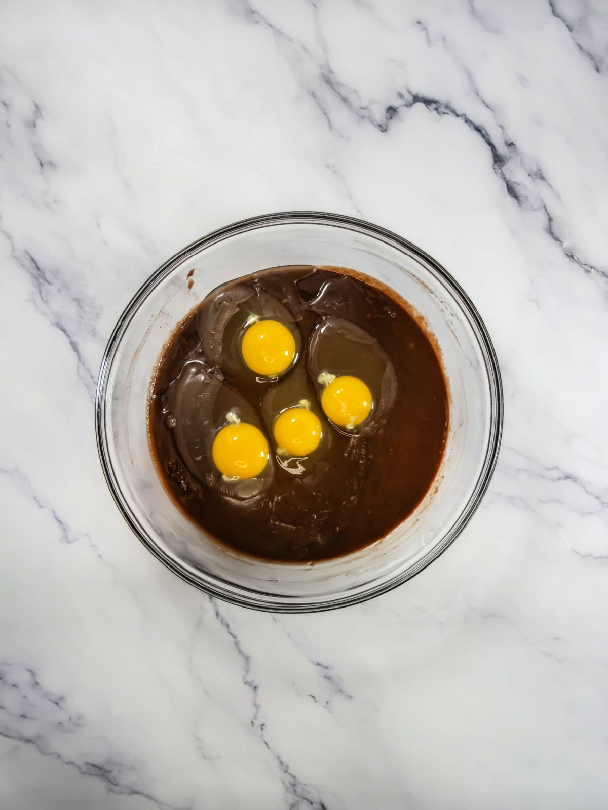 A glass bowl with brownie batter and four raw eggs, placed on a white marble countertop.