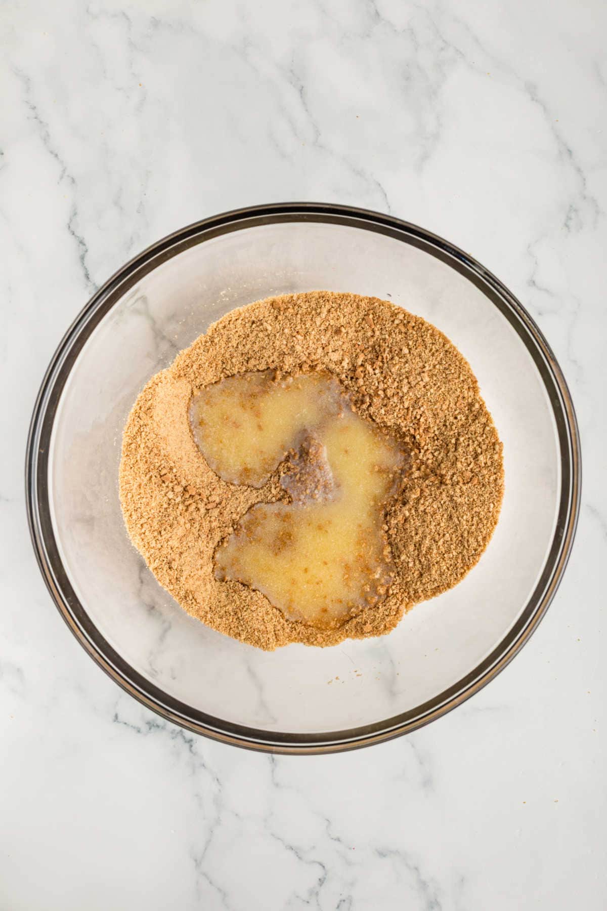 A glass bowl containing graham cracker crumbs and melted butter on a white marble surface.