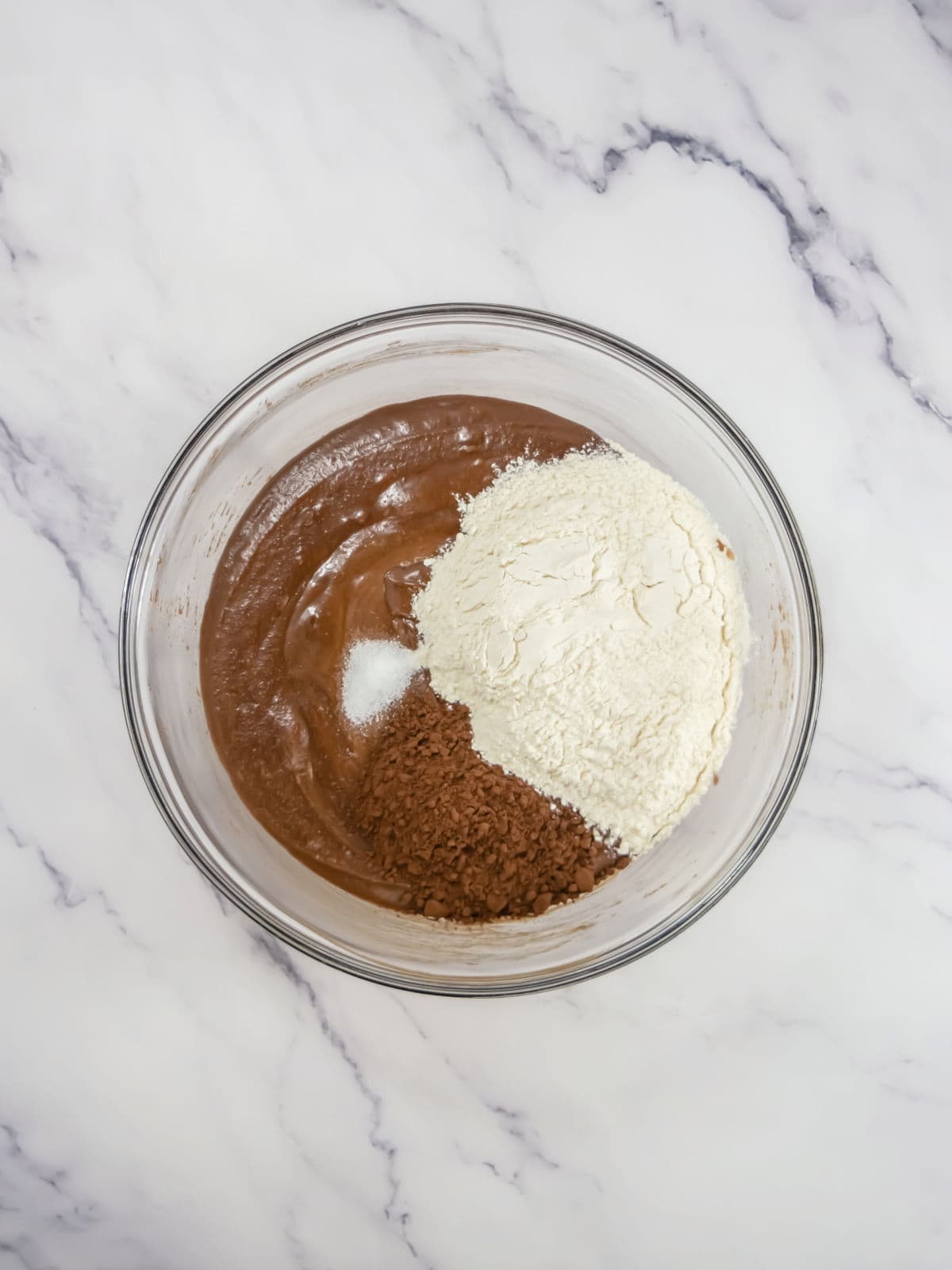 A glass bowl contains chocolate batter, flour, cocoa powder, and a small mound of salt on a white marble surface.