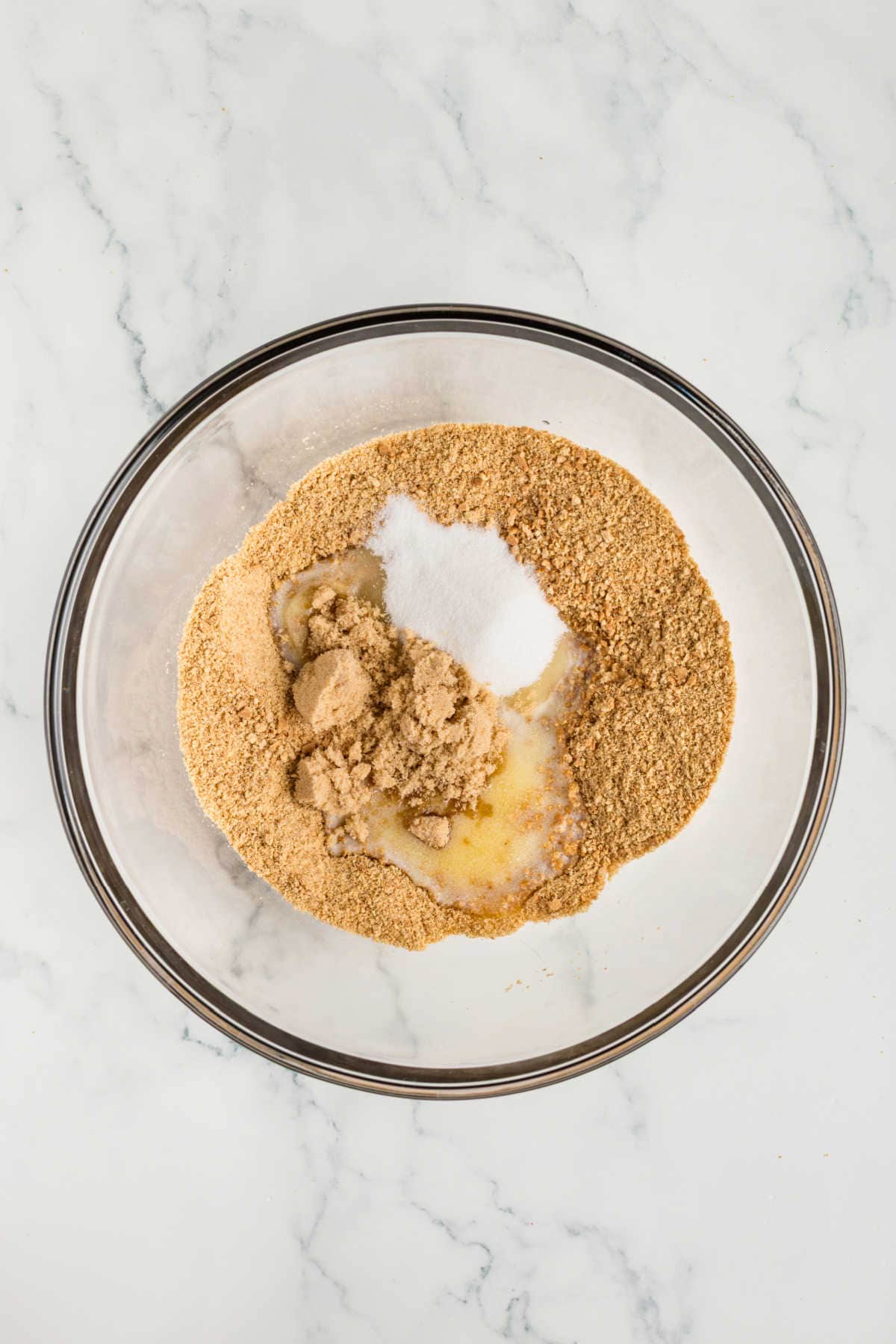 A glass bowl containing graham cracker crumbs, brown sugar, granulated sugar, and melted butter on a marble surface.