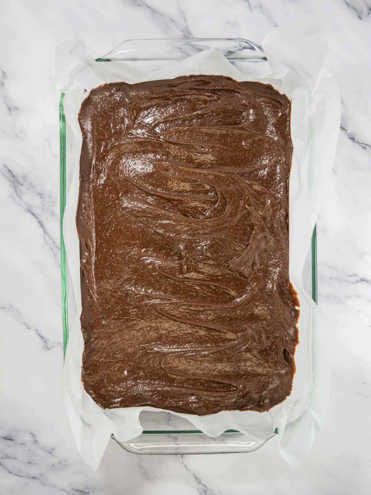 A glass baking dish lined with parchment paper, filled with unbaked chocolate brownie batter, sits on a white marble surface.