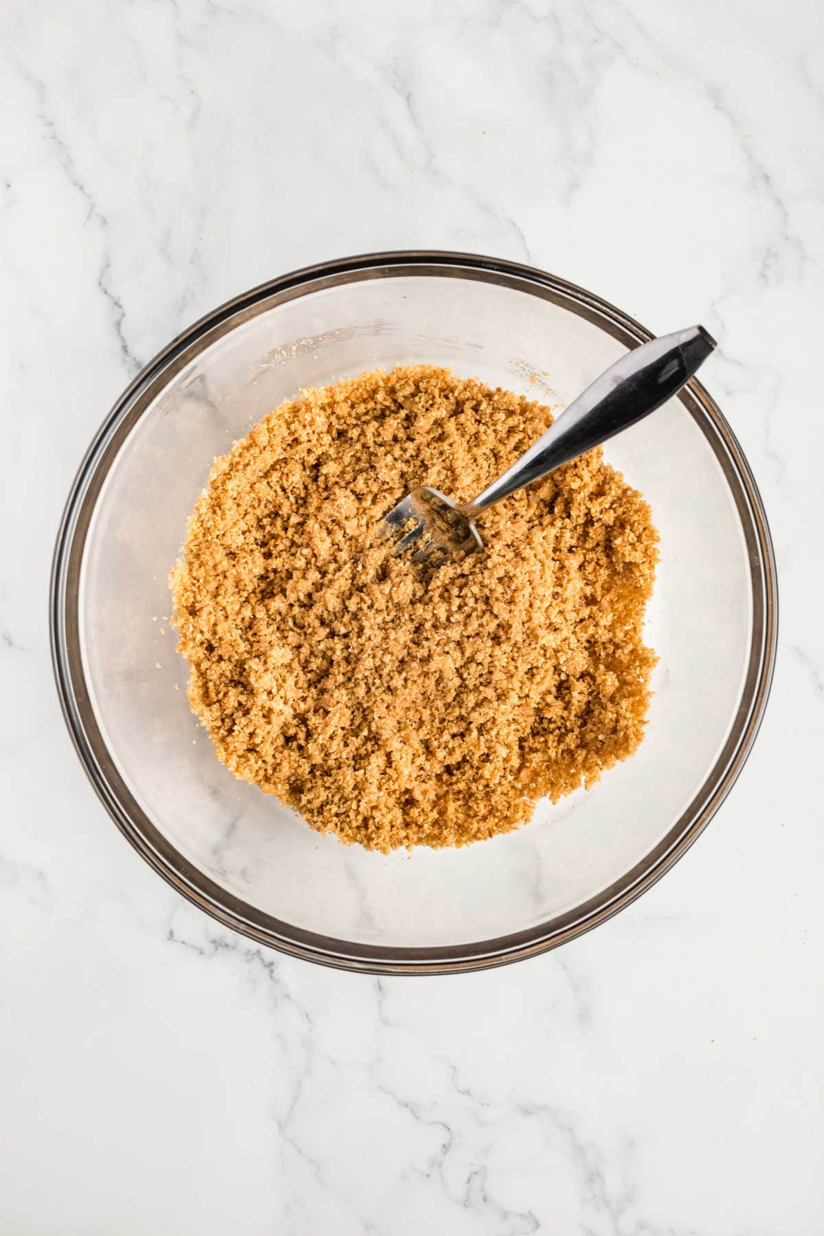 A clear glass bowl filled with crumbly brown mixture and a fork, placed on a white marble surface.