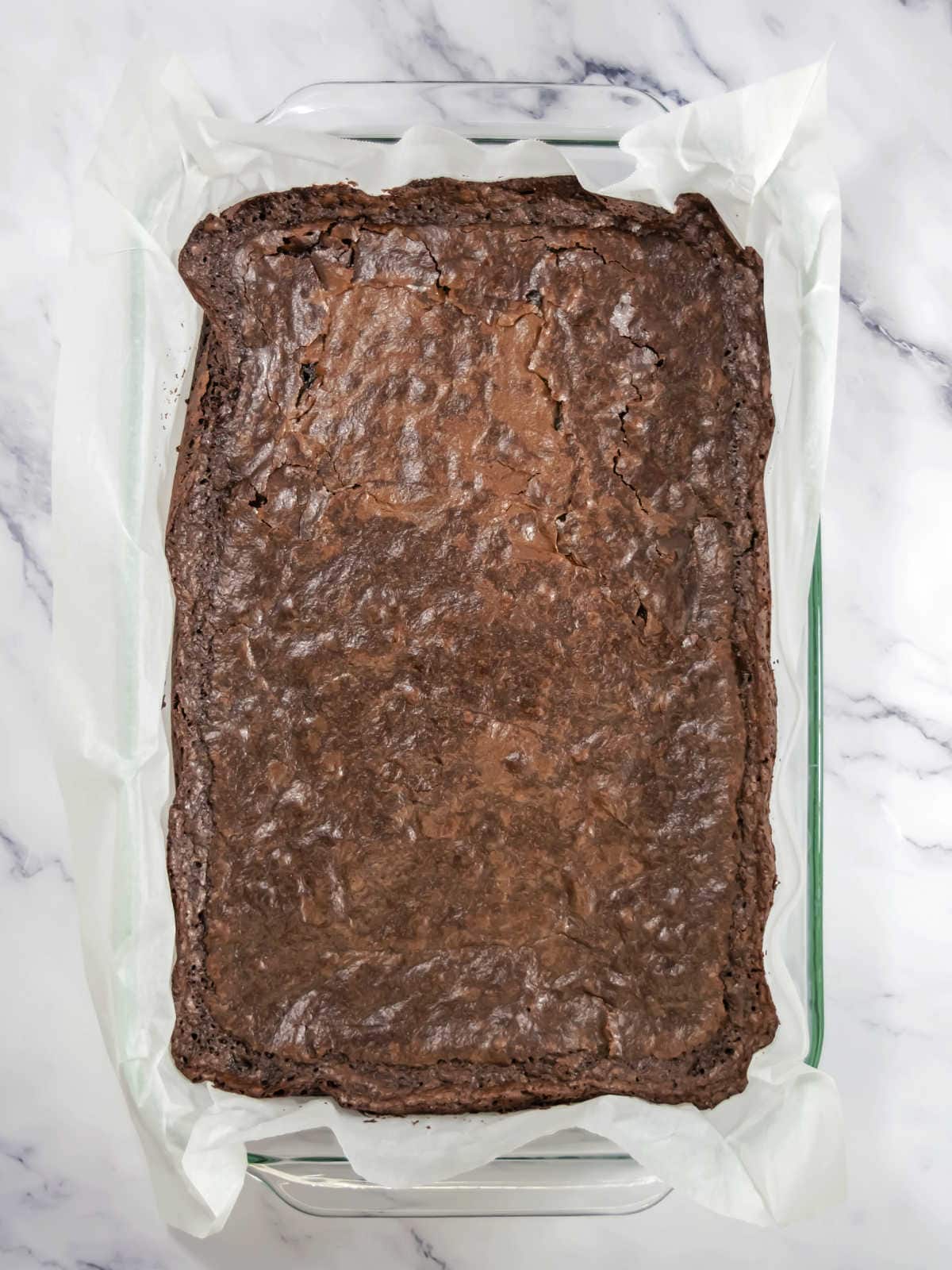 A rectangular pan lined with parchment paper containing a batch of baked brownies, shown from above on a marble surface.