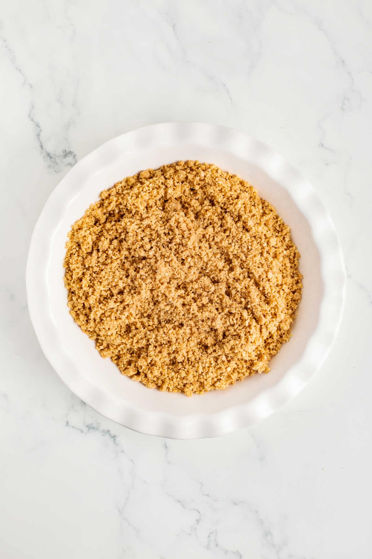 A white pie dish filled with an even layer of graham cracker crust mixture on a marble surface.