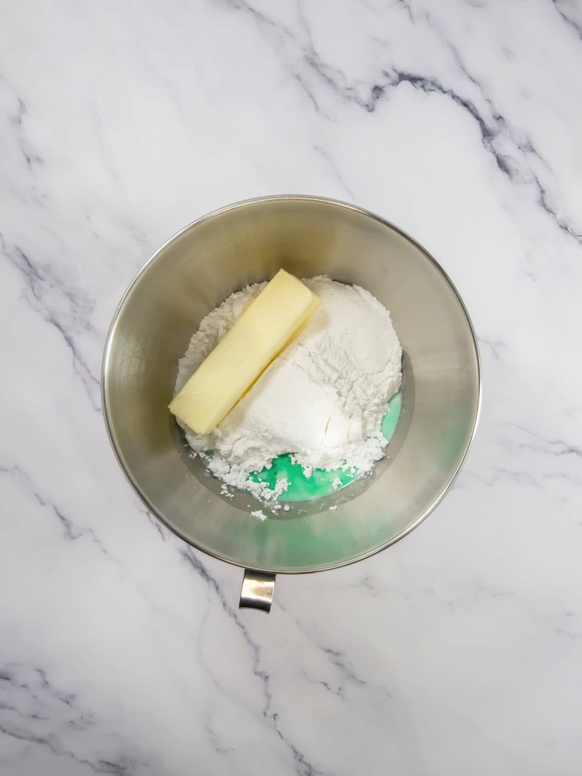 A stick of butter and a mound of flour in a stainless steel mixing bowl on a white marble surface.