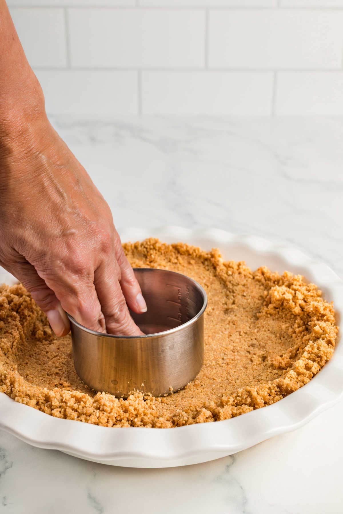 A hand presses a metal measuring cup into graham cracker crumbs in a pie dish to form a crust on a marble countertop.