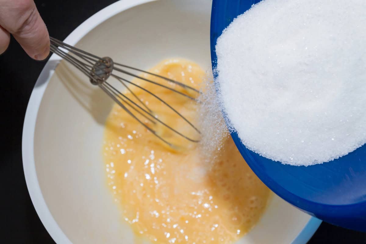 A person pours sugar from a blue bowl into a white bowl of beaten eggs while mixing with a whisk.