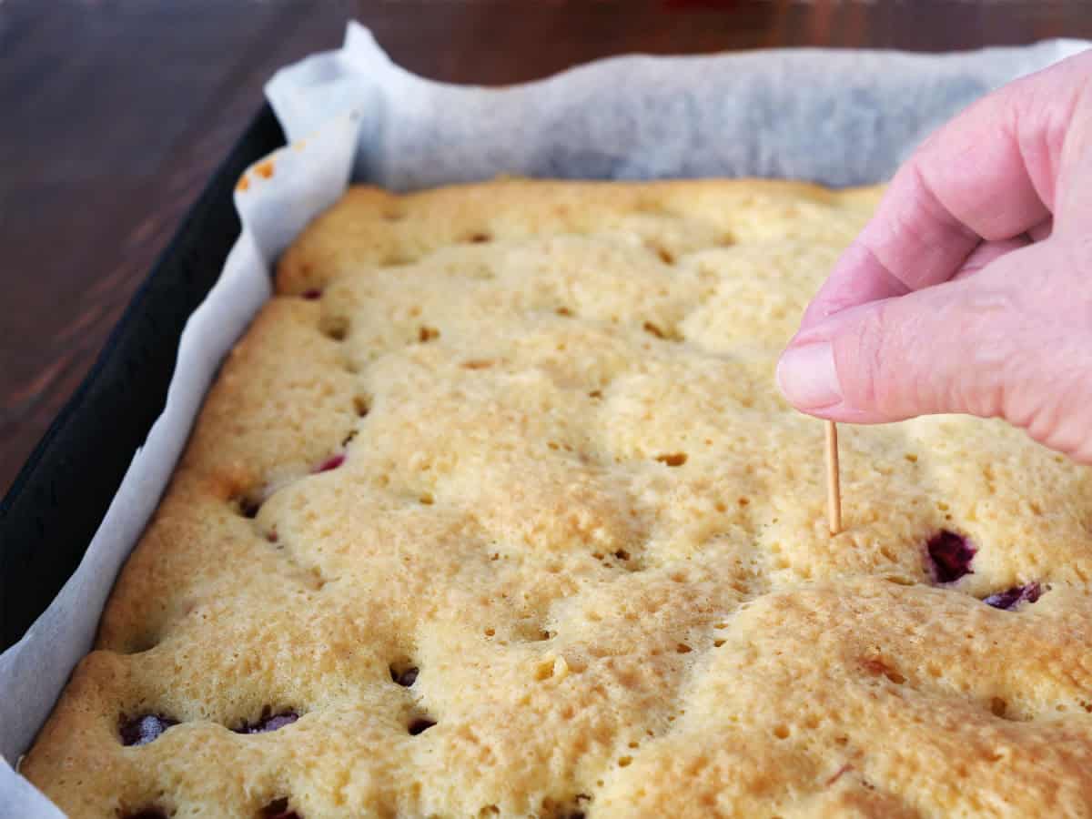A hand inserts a toothpick into a baked cake in a rectangular pan lined with parchment paper to check if it is fully cooked.