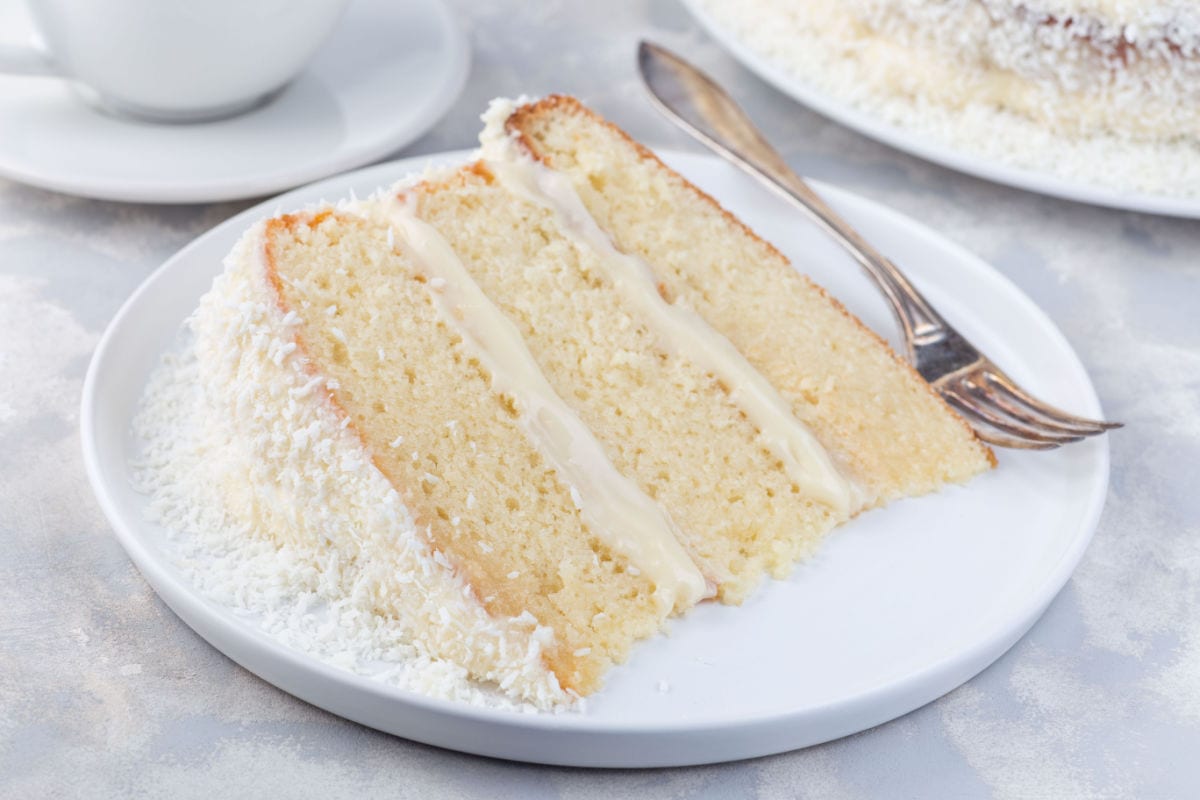 A slice of white cake with three layers and cream filling, coated with shredded coconut, served on a white plate with a fork.
