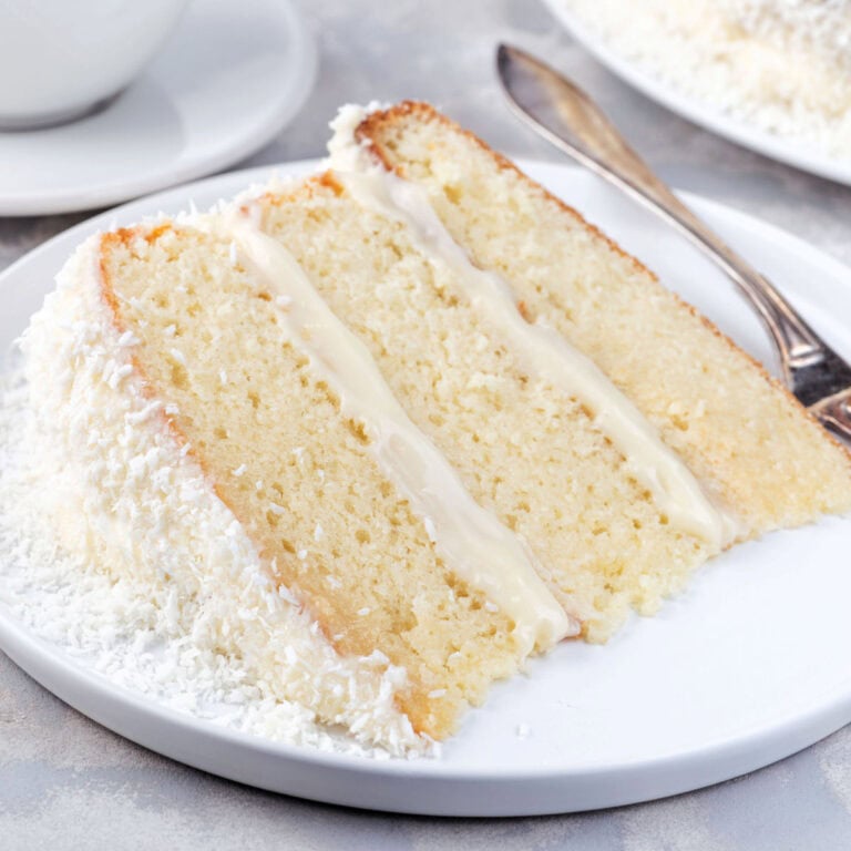 A slice of three-layer white cake with cream filling and shredded coconut on a white plate, with a fork next to it.