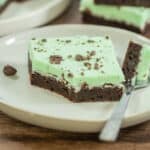A slice of brownie with a layer of green mint frosting and chocolate pieces on top, served on a white plate with a fork.
