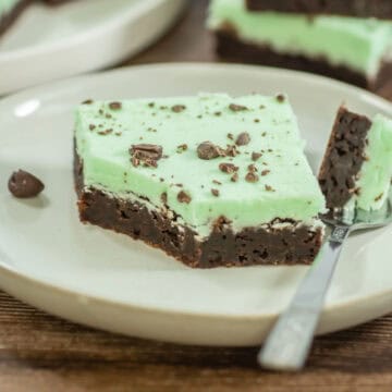 A slice of brownie with a layer of green mint frosting and chocolate pieces on top, served on a white plate with a fork.