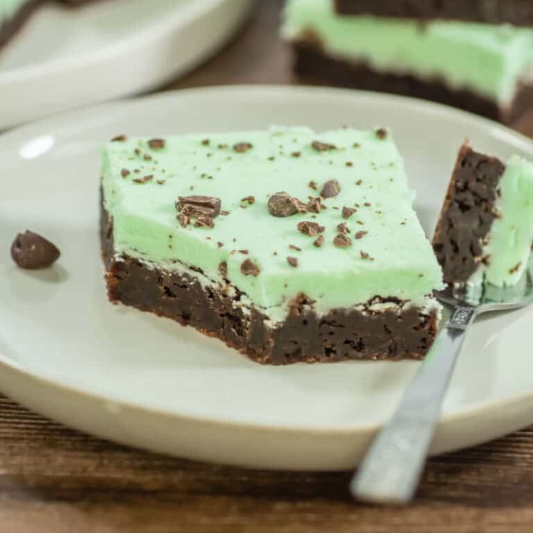 A slice of brownie with a layer of green mint frosting and chocolate pieces on top, served on a white plate with a fork.