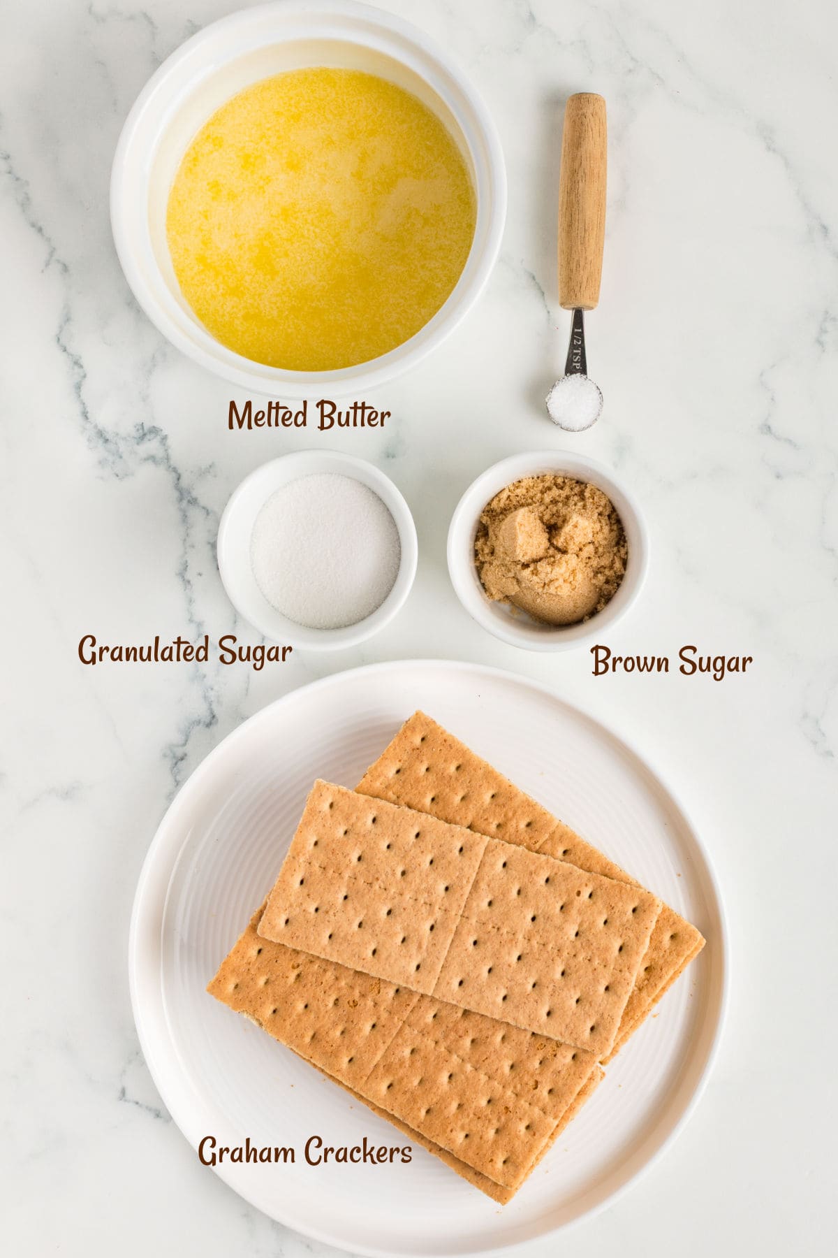 A plate with graham crackers, a bowl of melted butter, small bowls of granulated sugar and brown sugar, and a small spoonful of salt on a white surface.