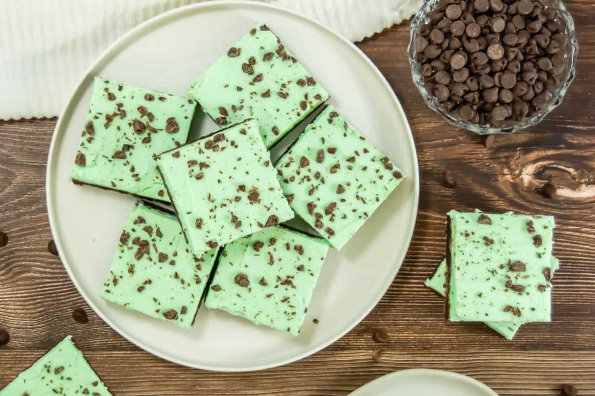 A plate of square, green mint brownies with chocolate chips on top, placed on a wooden table beside a bowl of chocolate chips.