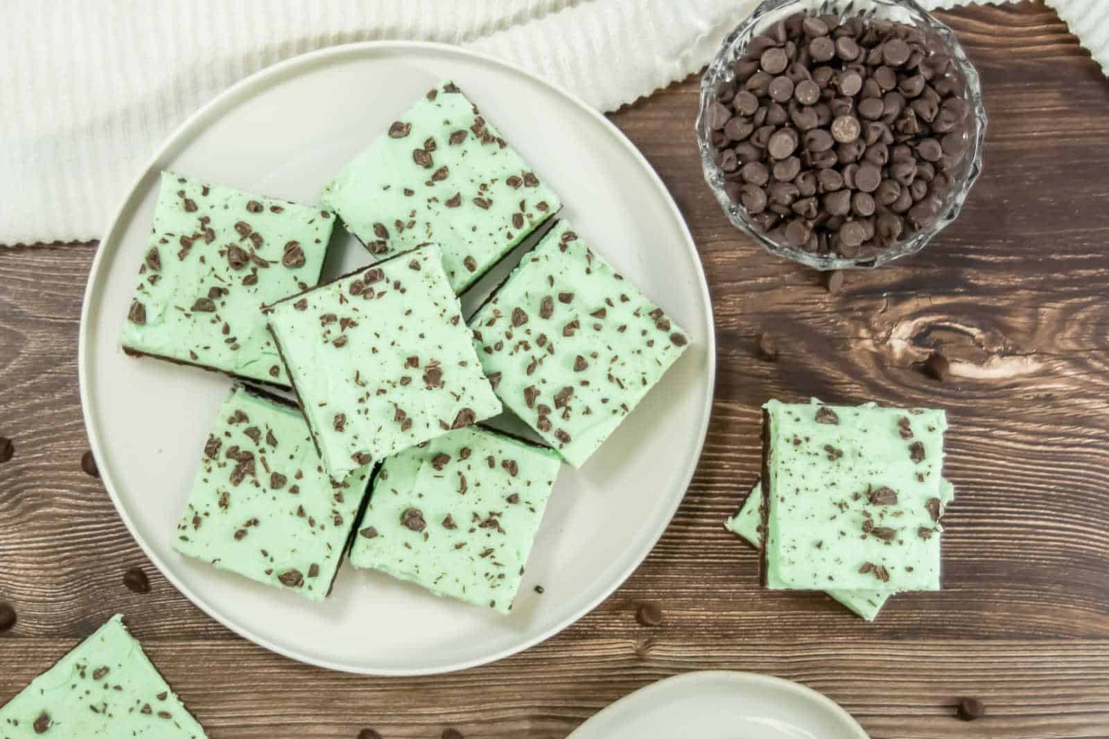 A plate of square mint chocolate chip fudge pieces sits on a wooden table next to a bowl of chocolate chips.