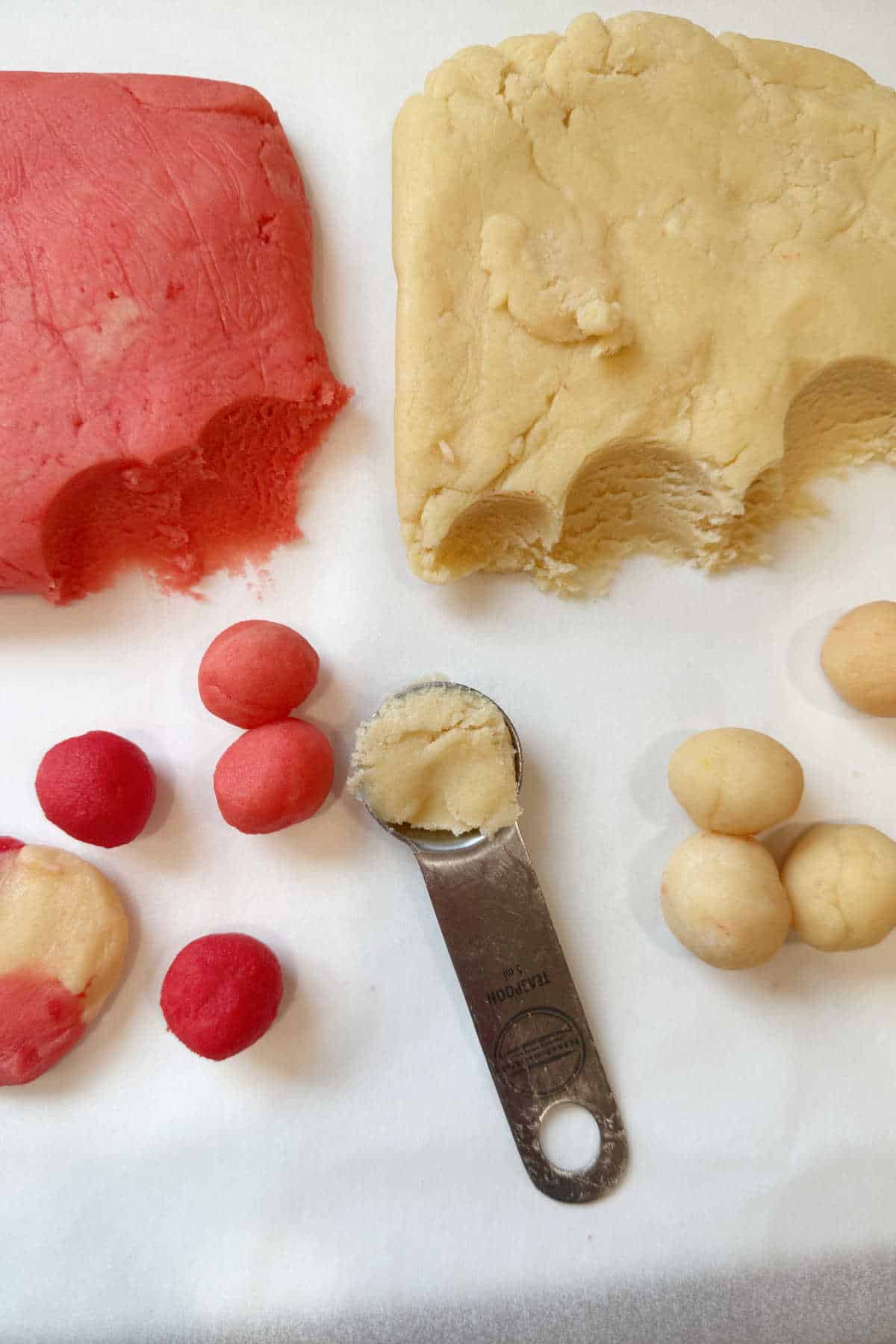 Two blocks of cookie dough, one red and one plain, sit on a surface with small dough balls and a tablespoon partially filled with plain dough nearby.
