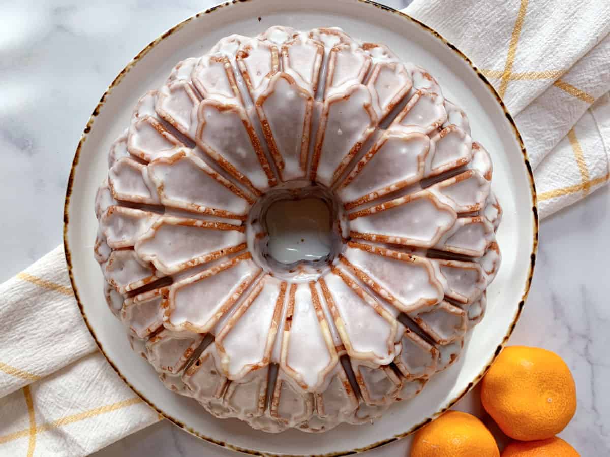 A glazed mandarin orange bundt cake on a white plate, with a white and yellow towel and three oranges beside it on a marble surface.