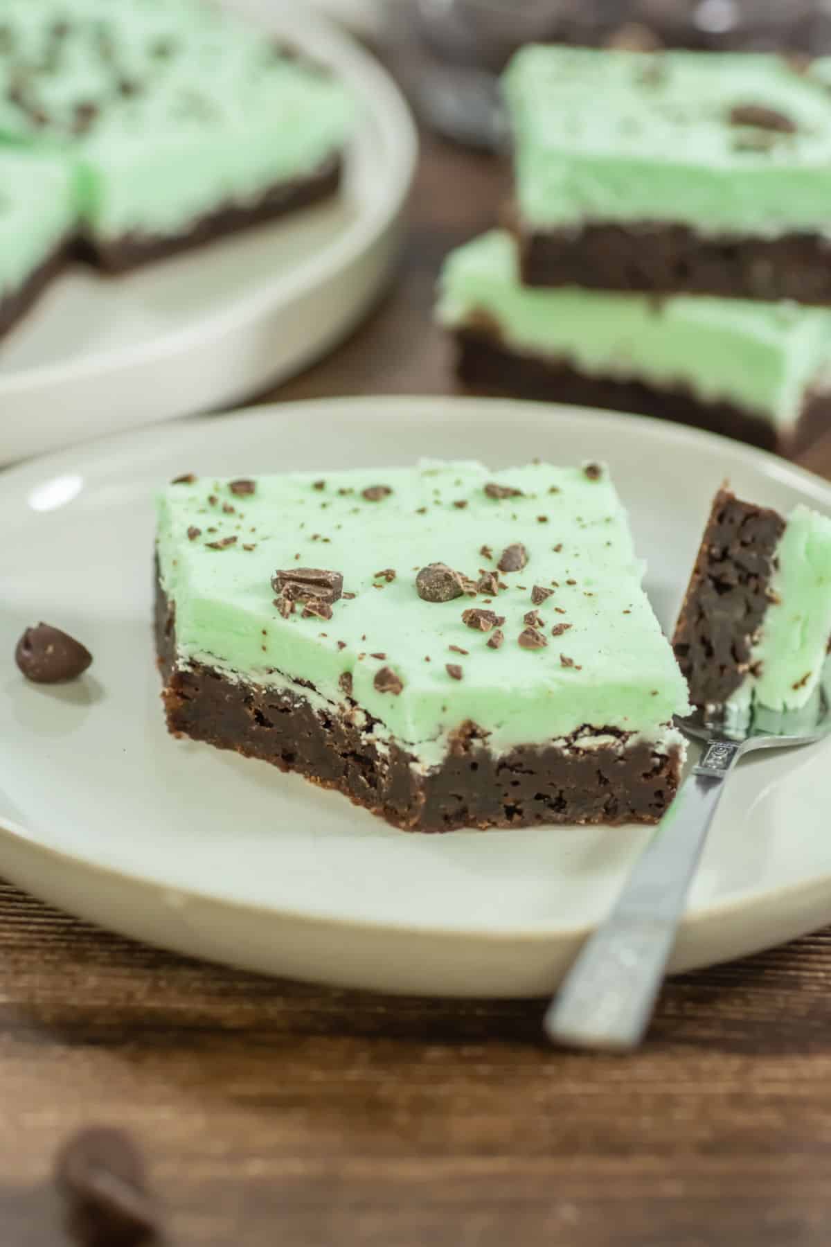 A square piece of chocolate brownie topped with green mint frosting and chocolate crumbles sits on a plate with a fork.